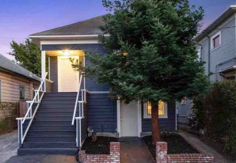 The two-story duplex features a blue exterior with white accents. A large tree stands in the front yard, providing some greenery. Light emanates from the entrance, highlighting the inviting stairs that lead to the upper unit.