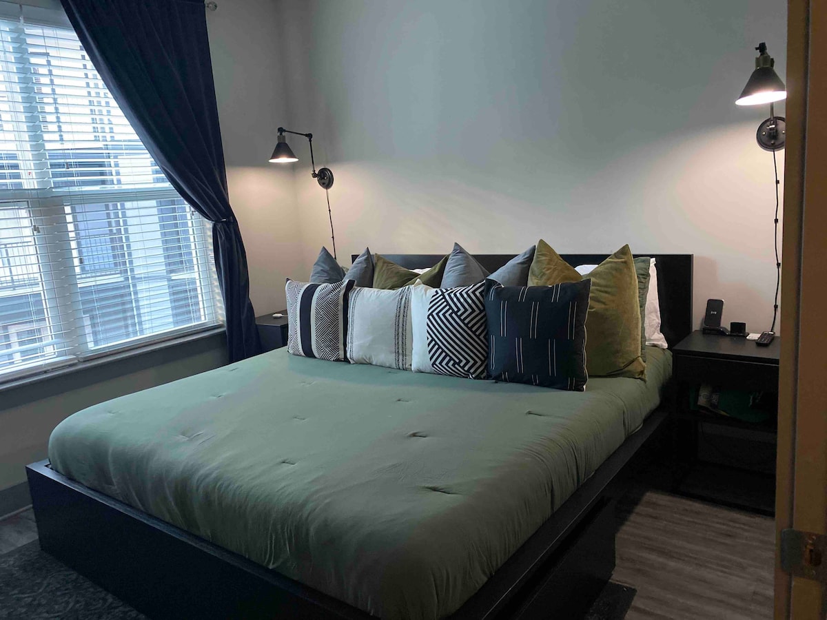 A spacious bed is adorned with a variety of decorative pillows in different textures and colors. Two wall-mounted lamps provide soft lighting beside the bed, and a large window with multiple panes allows natural light to fill the room, complemented by dark curtains.