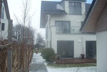 A modern two-story home is positioned among greenery, with large windows allowing natural light to fill the space. A wooden deck extends outward, bordered by potted plants. A clear pathway leads through the garden, enhancing the peaceful setting.