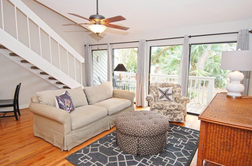 A bright living area offers a comfortable sofa and accent chairs with a patterned throw, all situated on a textured rug. Large glass doors bring in natural light and provide access to a deck with views of surrounding greenery.