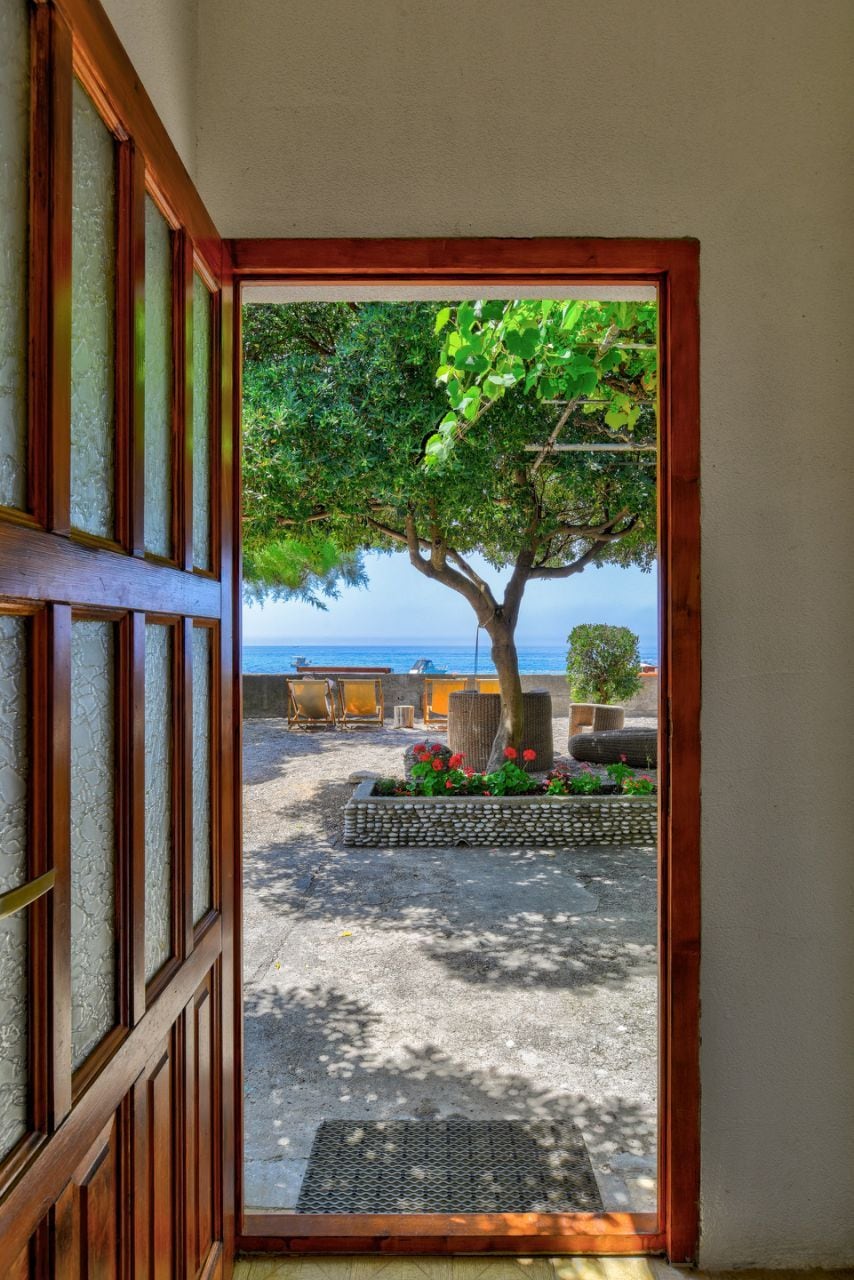 A view of a garden is framed by an open wooden door. A shaded tree stands in the center, surrounded by colorful flowers and plush lounge chairs. The distant beach can be seen, creating a serene outdoor space.