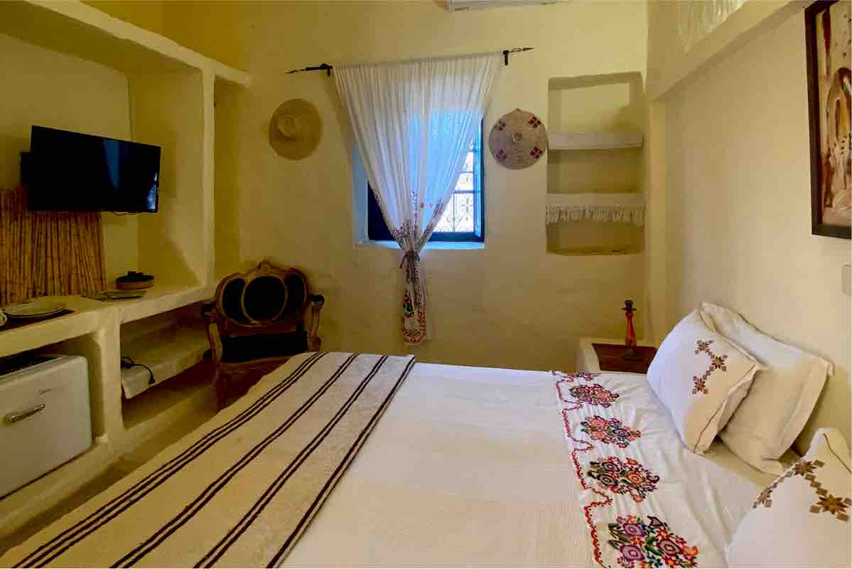 A cozy room features a spacious bed adorned with decorative pillows and an embroidered blanket. A small table with a decorative item sits beside the bed. A television is mounted on the wall, and a window is dressed with light curtains, allowing natural light to filter in.