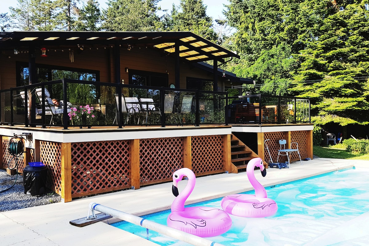 A heated pool is framed by a wooden deck, featuring two pink flamingo floaties resting on the water's surface. The adjoining house showcases a covered patio furnished with dining tables and chairs, surrounded by lush greenery.