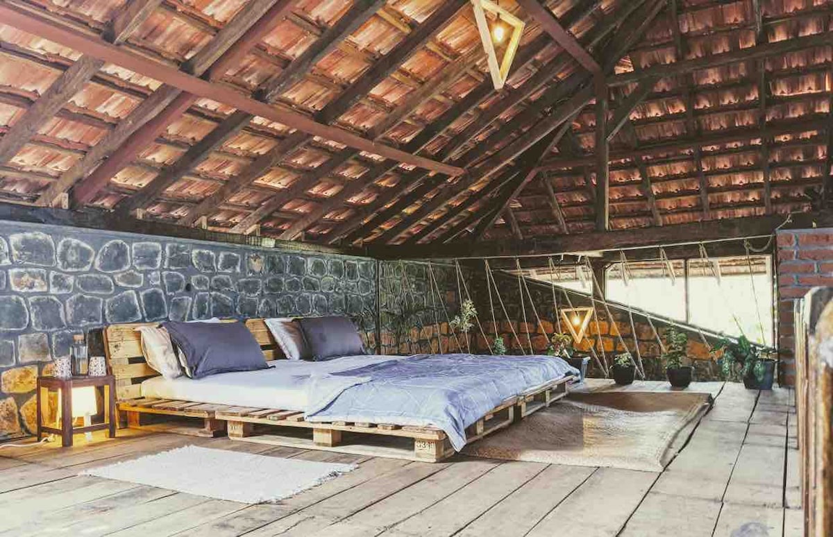 An attic space features a spacious sleeping area with a low wooden pallet bed topped with soft bedding and pillows. Natural light filters through a window, while the wooden ceiling and walls create a rustic ambiance. A small lantern adds a touch of warmth.