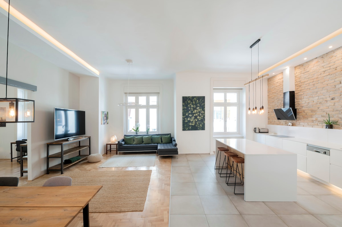 A spacious combined kitchen-living room features a modern kitchen island with stools, a full-sized refrigerator, and sleek cabinetry. A large sofa is positioned near a 75" TV, while natural light fills the space through multiple windows, enhancing the warm tones of the decor.