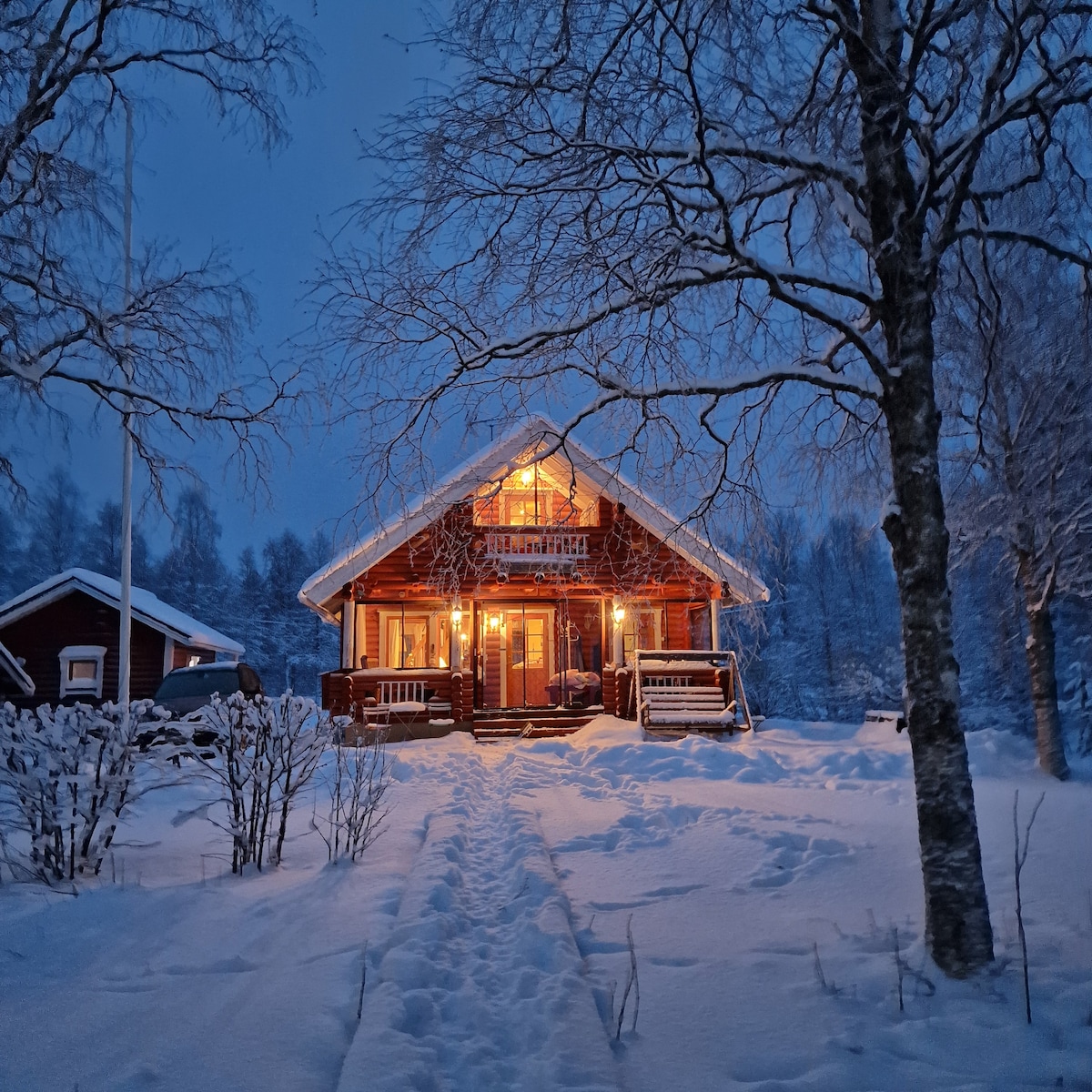 A two-story cabin is surrounded by snow-covered landscapes, its warm lighting glowing through large windows. The entrance features a staircase leading to a covered porch, framed by trees adorned with snow. A clear path leads towards the inviting front door.