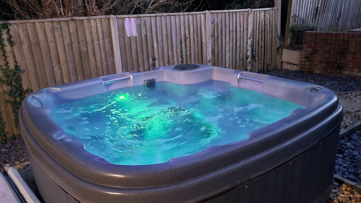 A spacious hot tub is illuminated with a soft blue and green glow, gently bubbling with water. Surrounded by a wooden fence, the area features natural stone pathways and is positioned within a serene garden setting.