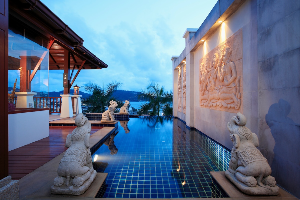 A private pool features elegant stone sculptures, adding a touch of Thai architecture. The serene water reflects the night sky, while the surrounding area is illuminated, creating a peaceful ambiance. Lush greenery can be seen in the background, enhancing the villa's natural beauty.