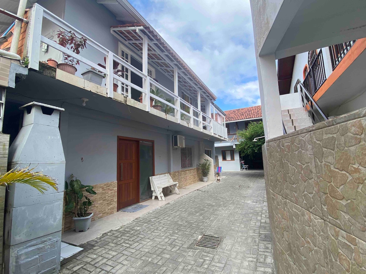 A spacious courtyard is framed by two buildings, showcasing a mix of outdoor seating options and greenery. A white wooden bench is positioned on one side, while nearby, a stone wall provides texture to the layout. The sky features soft clouds, adding to the serene atmosphere.