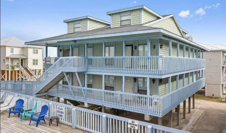 A two-story building features a light blue exterior with multiple balconies. A wide staircase leads to the second level. In the foreground, several blue lounge chairs are arranged near the entrance, creating a welcoming outdoor space.
