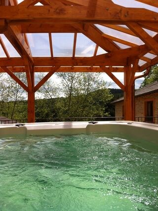 A wooden gazebo provides shelter over a relaxing hot tub, surrounded by lush green trees and a scenic view. The water appears inviting, glistening under the natural light that filters through the open roof, creating a tranquil atmosphere.