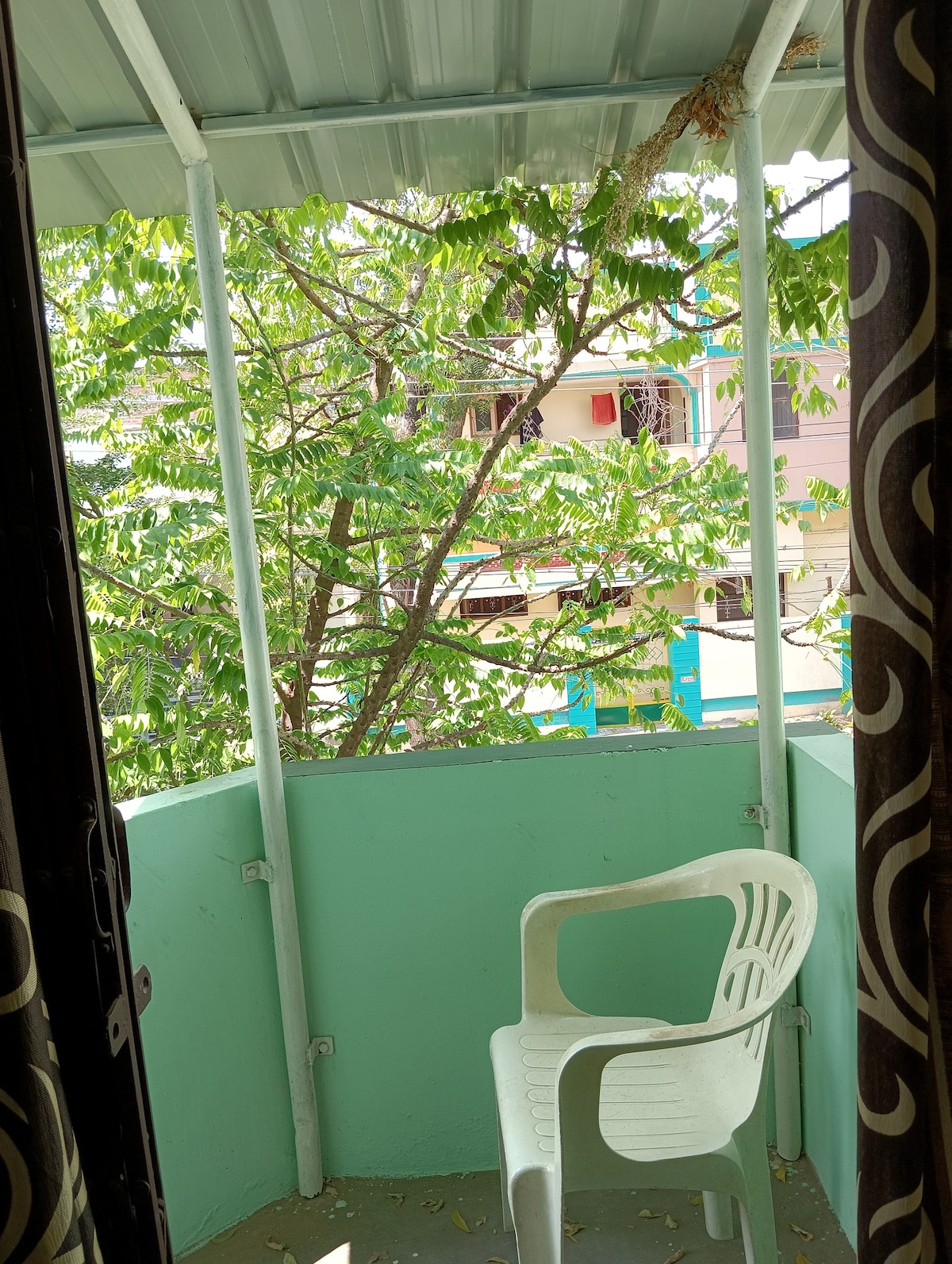 A private balcony space is visible, featuring a white chair positioned against a pale green wall. Lush green foliage from the adjacent tree offers a sense of serenity, while colorful buildings can be seen in the background, enhancing the outdoor ambiance.