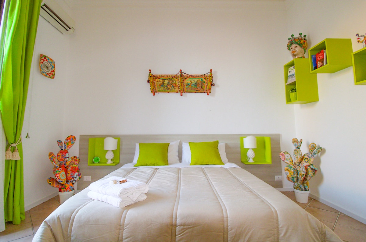 A bright bedroom features a spacious bed with neutral linens and vibrant green accent pillows. Colorful decor, including wall art and potted plants, enhances the inviting atmosphere. Natural light enters through a window with sheer curtains, complementing the white walls and soft floor tiles.