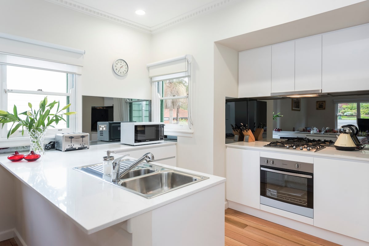 The kitchen features modern white cabinetry and sleek countertops. A double sink is positioned in front of a window, while essential appliances, including a microwave and gas stove, are neatly arranged. Natural light illuminates the space, creating a bright and functional cooking area.
