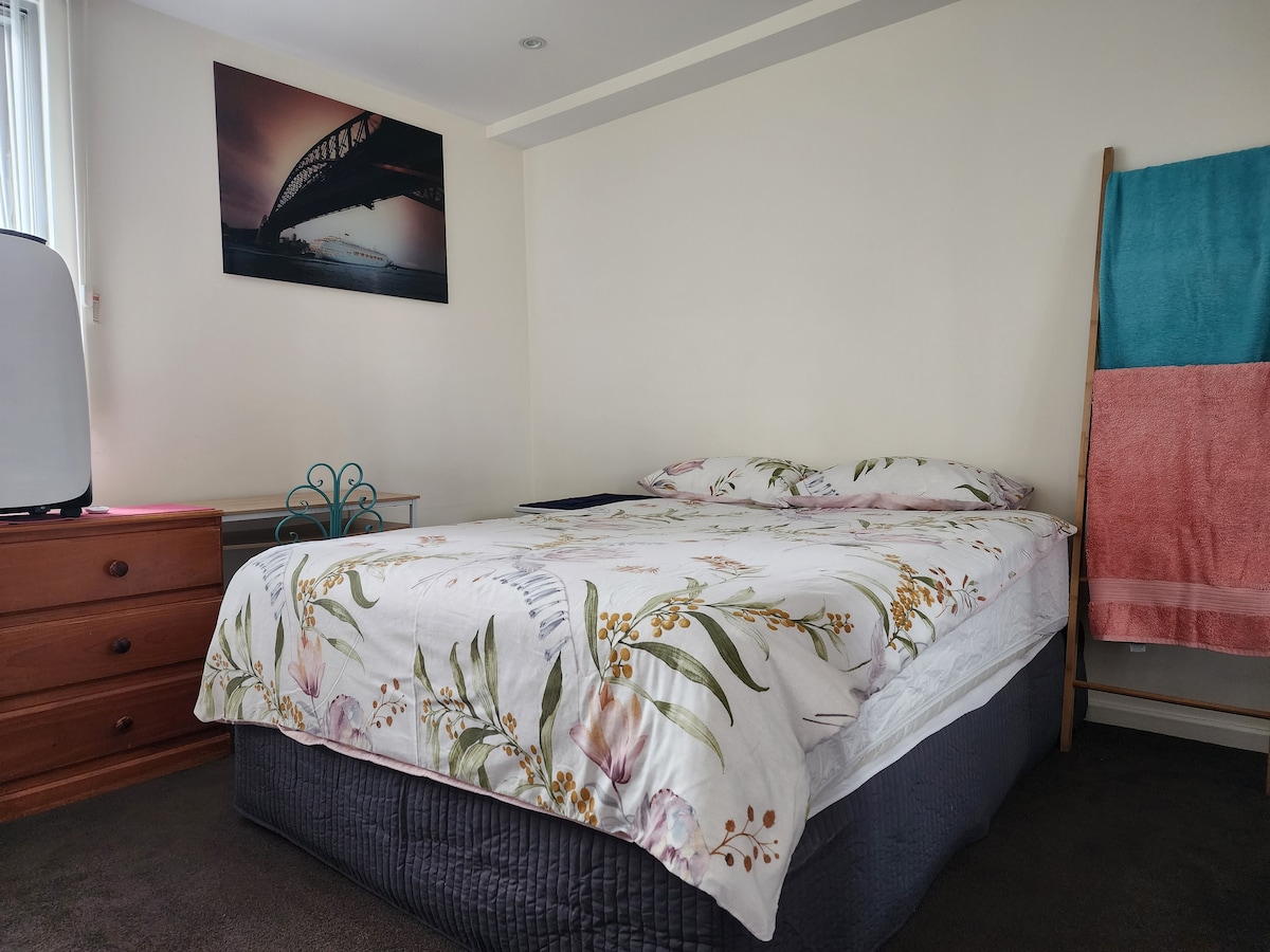 A private room features a spacious bed dressed in floral linens, complemented by a wooden dresser and a turquoise and pink towel rack. Natural light filters in through a window, and a wall art piece displays a bridge over water.
