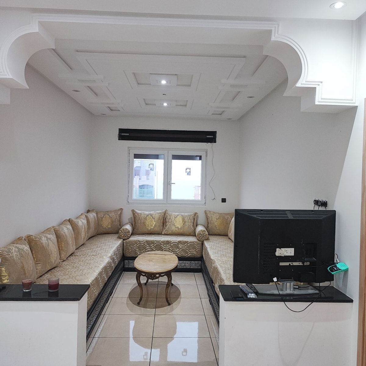 A well-lit living area features a spacious corner seating arrangement with decorative cushions. A round wooden coffee table rests at the center, while a television is positioned on a modern stand. Large windows allow natural light to illuminate the room.