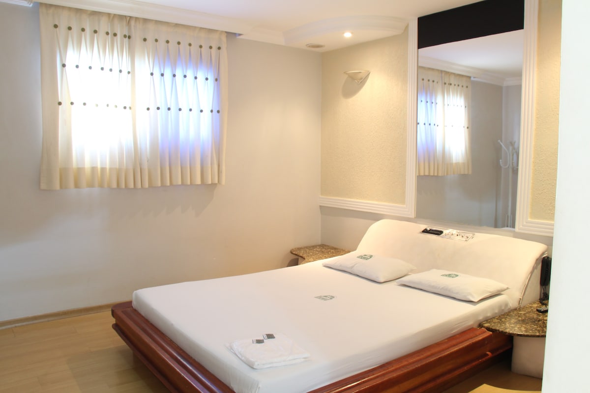 A spacious room features a large bed with neatly arranged pillows. Natural light filters through two windows, dressed with soft drapes. A mirrored wall contributes to an open feel, while a small bedside table adds functionality. Warm tones create a calming ambience.