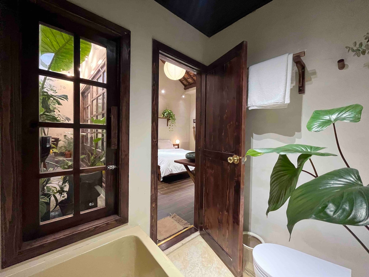 A doorway opens to a cozy space, revealing a well-lit bedroom adorned with plants. A large window allows natural light to flow in, complementing the earthy tones of the walls and decor. The inviting bathroom features a tub and a toilet beside a leafy plant.