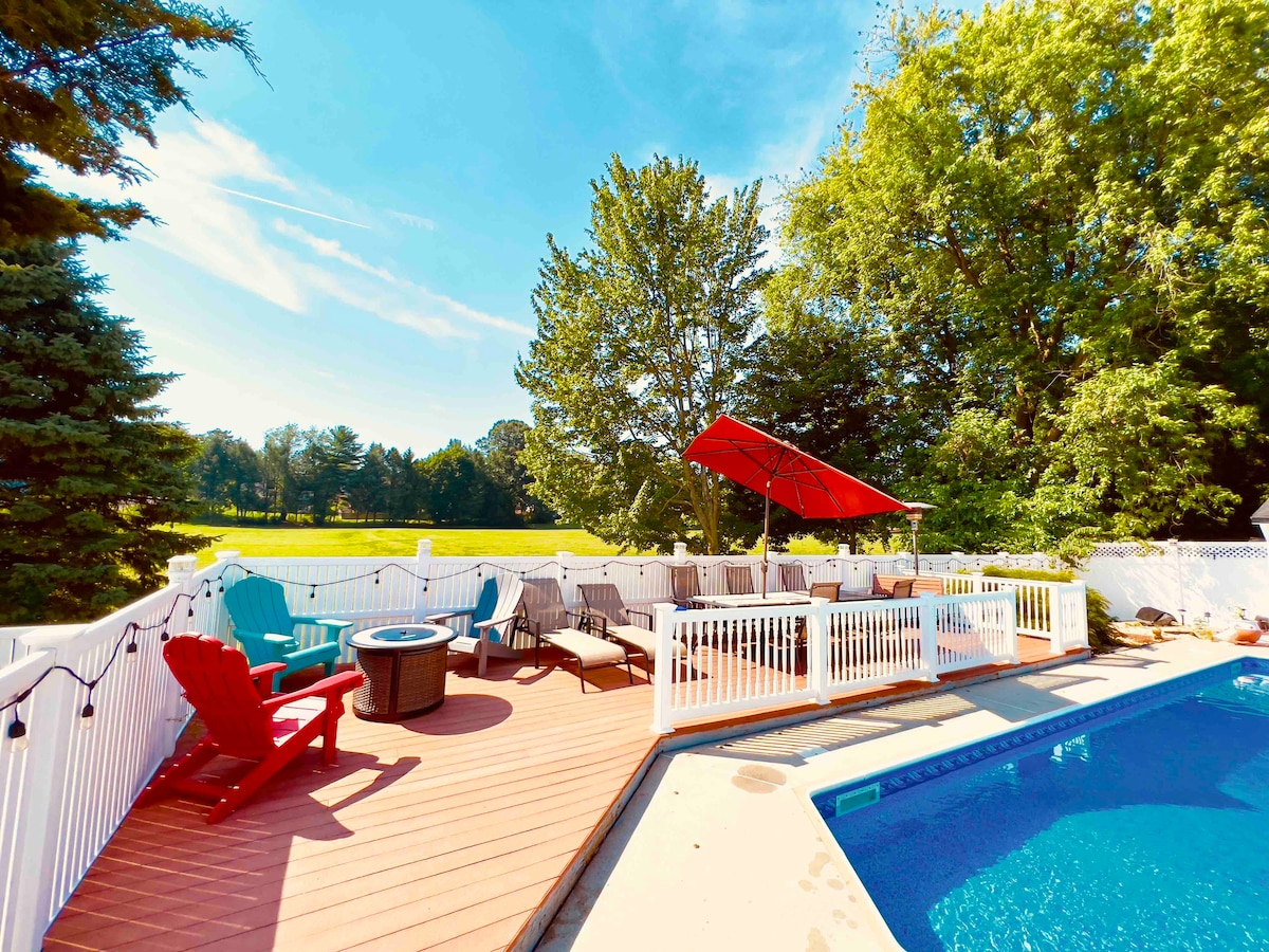 A spacious outdoor patio surrounds a vibrant blue pool, featuring lounge chairs and a fire pit. A red umbrella provides shade for seating. Lush greenery and open fields are visible in the background, creating a serene environment for relaxation.