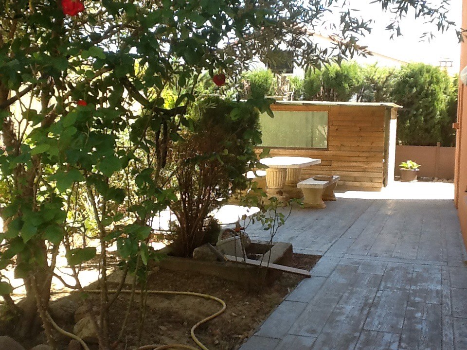 A wooden terrace is visible, surrounded by greenery and flowering plants. A rustic wooden structure with windows is positioned in the background. A table and two stone benches are placed on the terrace, providing a serene outdoor space for relaxation.