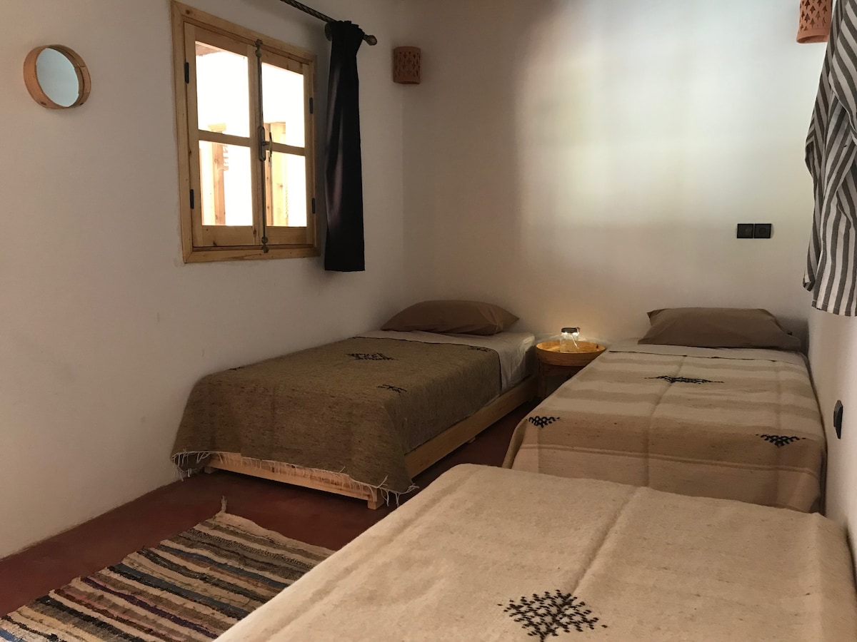 A simple and comfortable room is furnished with two spacious beds, each covered with traditional Berber blankets. A small side table with a lamp sits between them. Natural light filters through the window, which is framed by a dark curtain, enhancing the cozy atmosphere.