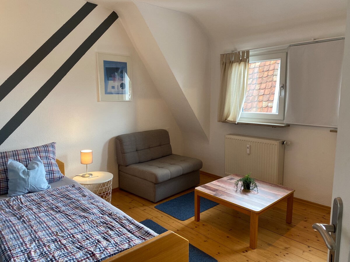 The room features a spacious bed with a patterned blanket and a cozy gray sofa. A small table is placed on a blue rug. Natural light enters through a window, enhancing the clean, simple design highlighted by a decorative framed picture.