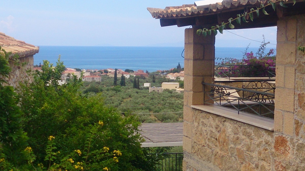 A view of the serene sea is framed by an open veranda, showcasing stone architecture and lush green foliage. The landscape features rolling hills and scattered traditional homes beneath a clear sky, creating a peaceful setting.
