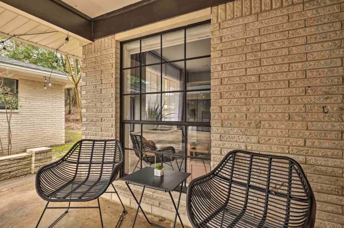 A small outdoor seating area is presented with two black wire chairs and a matching side table. Behind the seating, a large window offers a view into a light-filled interior space. The brick wall adds texture and warmth to the cozy setting.