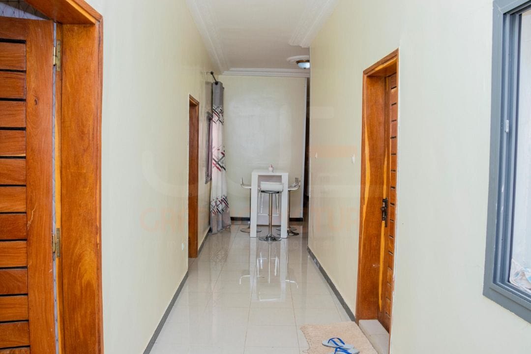 A well-lit hallway features smooth, polished flooring and neutral-toned walls. On one side, wooden doors provide access to rooms, while a small table with chairs is positioned in the center. Natural light enters through a window at the end of the corridor.
