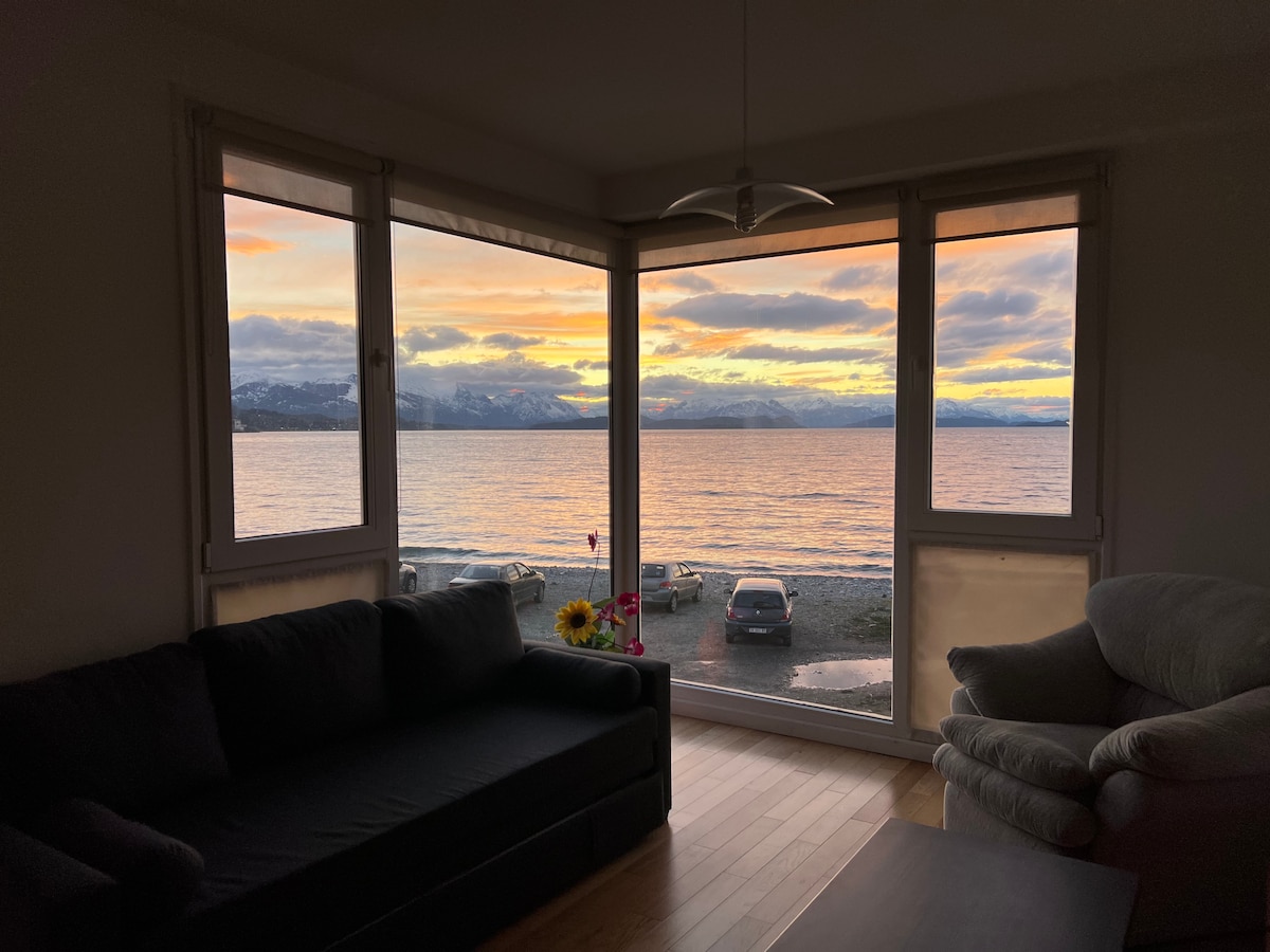 A living area is framed by large windows that overlook Lake Nahuel Huapi, showcasing a vibrant sunset. The scene features a sandy beach and distant snow-capped mountains. A black sofa and a light-colored armchair are positioned for relaxing while enjoying the view.