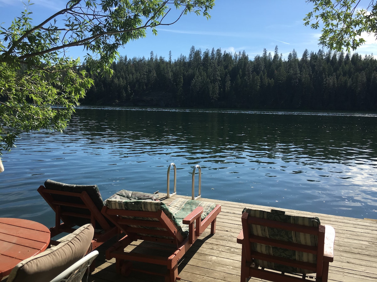 A private dock extends over calm waters, framed by lush trees. Two loungers with cushions are positioned for relaxation, alongside a round table. The serene lakeshore is visible, reflecting the vibrant blue sky and surrounding greenery.