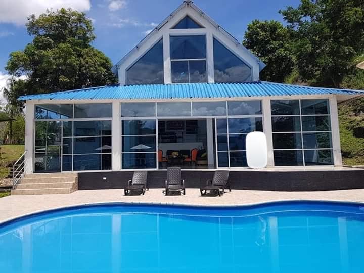 A modern house with a blue roof features large glass windows reflecting the surroundings. A spacious patio is seen in front of a refreshing pool. Lounge chairs are arranged near the pool, offering a relaxing space to enjoy the view.