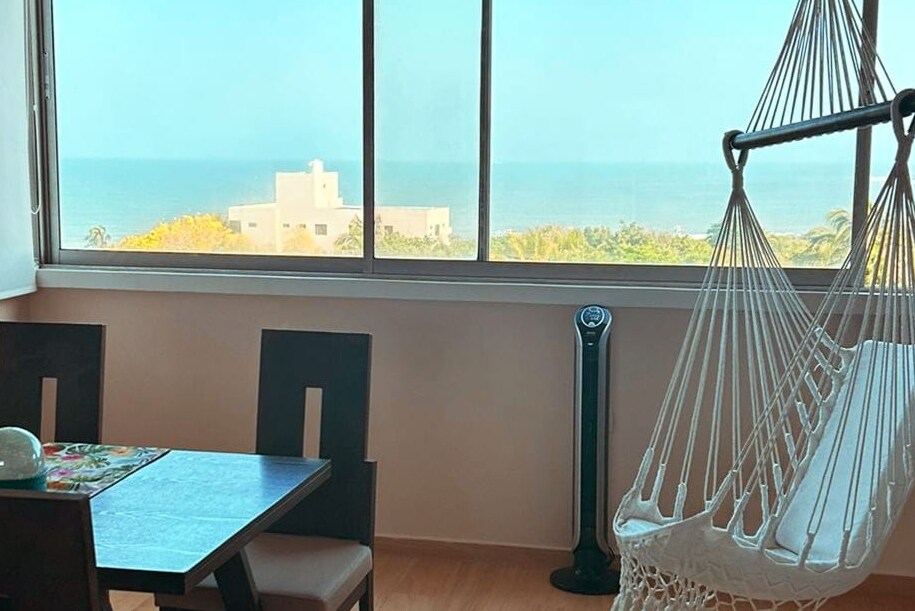 A dining area features a wooden table and two chairs, complemented by a hammock hanging near a large window. The ocean view is visible through the clear glass, creating a bright and airy atmosphere.