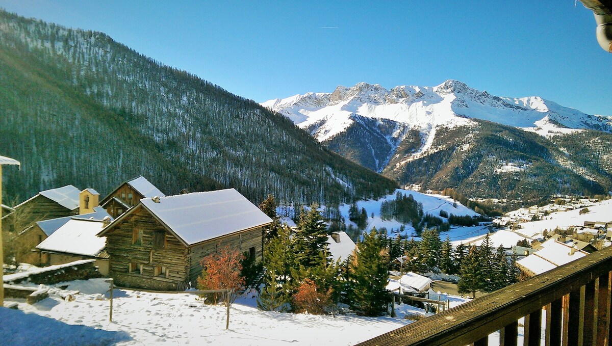 A scenic view highlights snow-covered mountains and a valley dotted with wooden chalets. In the foreground, a rustic stone chalet is partially visible, framed by evergreens against a clear blue sky.
