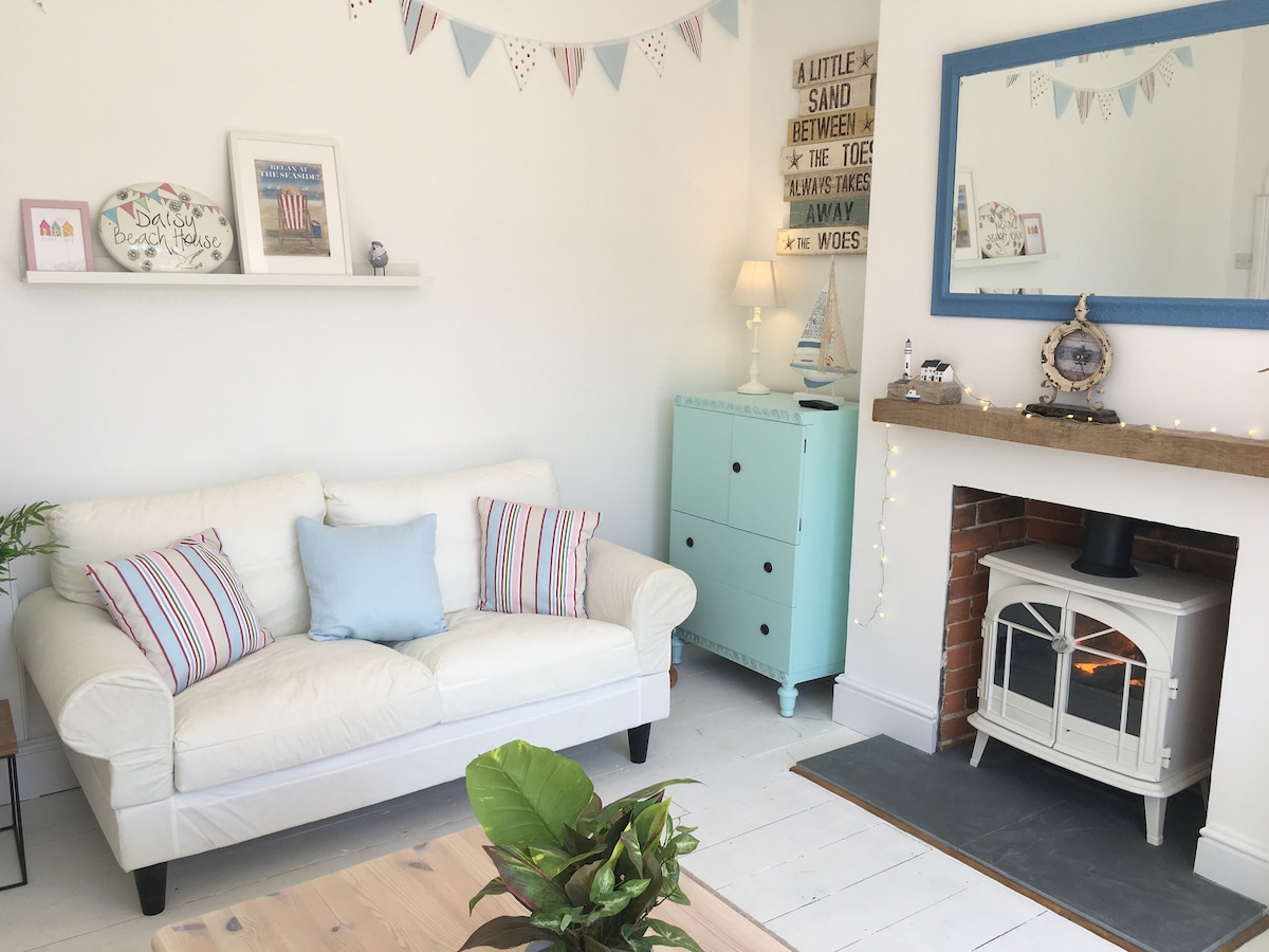 A bright sitting room features a white sofa adorned with colorful striped cushions, set against clean white walls. A blue-framed mirror complements a rustic mantel with fairy lights, while a decorative cupboard provides a pop of color. An electric log burner adds warmth to the space.