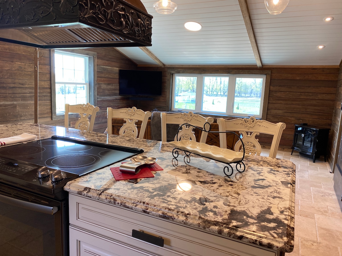 A spacious kitchen features a large island with intricate granite countertops and an integrated cooktop. Elegant dining chairs surround a rustic table, while large windows provide a view of the lavender field. Wood paneling and a flat-screen TV add warmth to the inviting atmosphere.