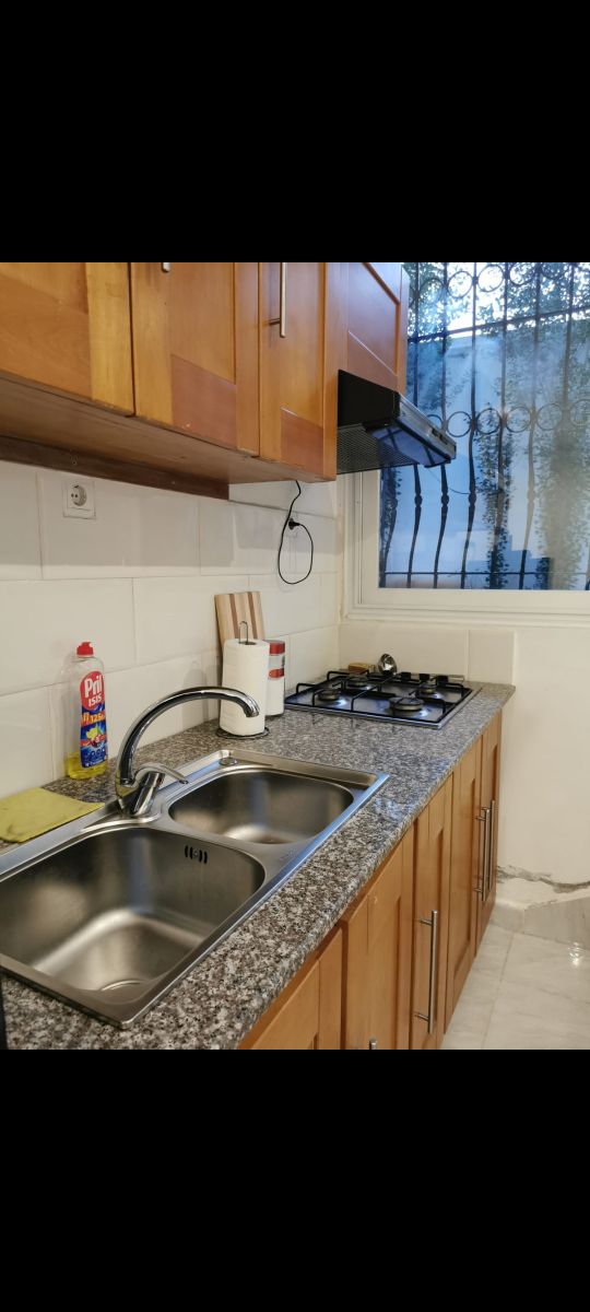 A compact kitchen features wooden cabinetry and granite countertops. The two stainless steel sinks are complemented by a gas stove with multiple burners, while a window allows natural light to brighten the space.