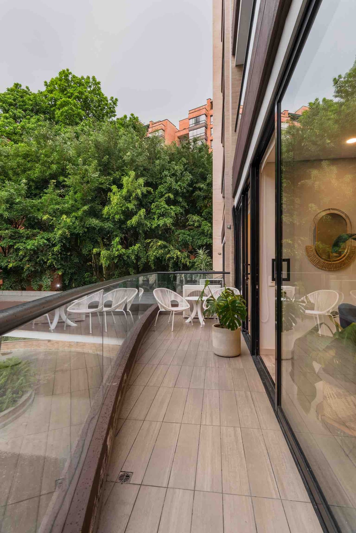 A spacious balcony is featured, surrounded by lush greenery. The space includes two white chairs and a potted plant, offering a peaceful retreat. Large glass doors provide easy access to the interior, emphasizing a seamless connection between indoor and outdoor living.