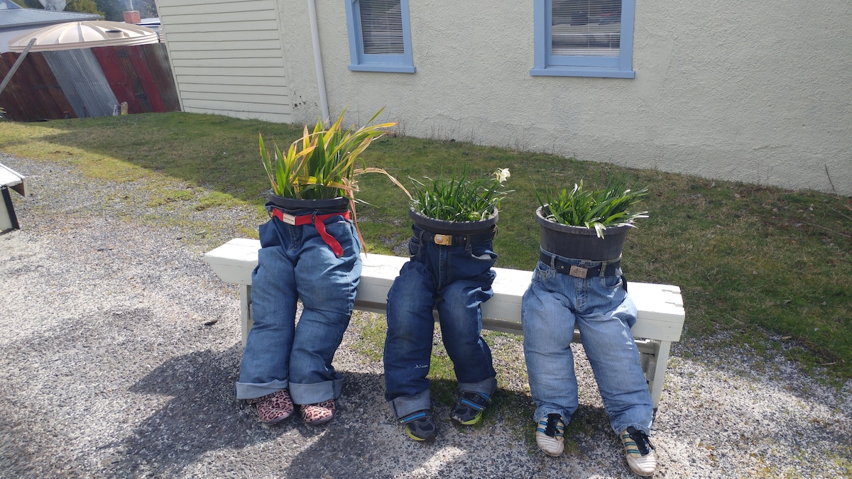 Three planters creatively arranged as human figures sit on a wooden bench. Each planter is made from denim jeans filled with greenery, featuring different footwear. The background includes a grassy area and a pale-colored structure with blue window frames.