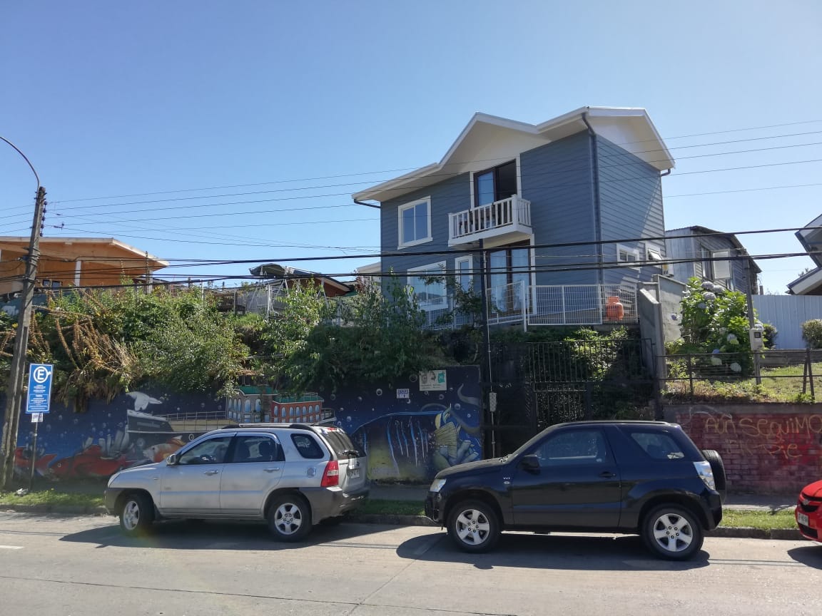 A two-story gray house is positioned on an elevated lot, featuring a small balcony on the upper level. The surrounding area includes a garden with greenery. Two parked cars are visible in front, and a colorful mural decorates the wall at the base of the property.