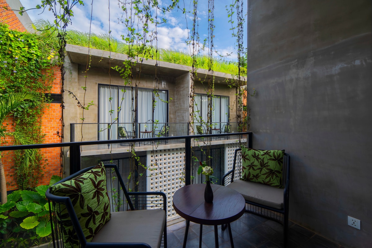 A small private terrace features two seating chairs adorned with leaf-patterned cushions, complemented by a round table. Lush greenery surrounds the area, with vines cascading from above. Large glass doors lead to the rooms, while a bright sky is visible overhead.