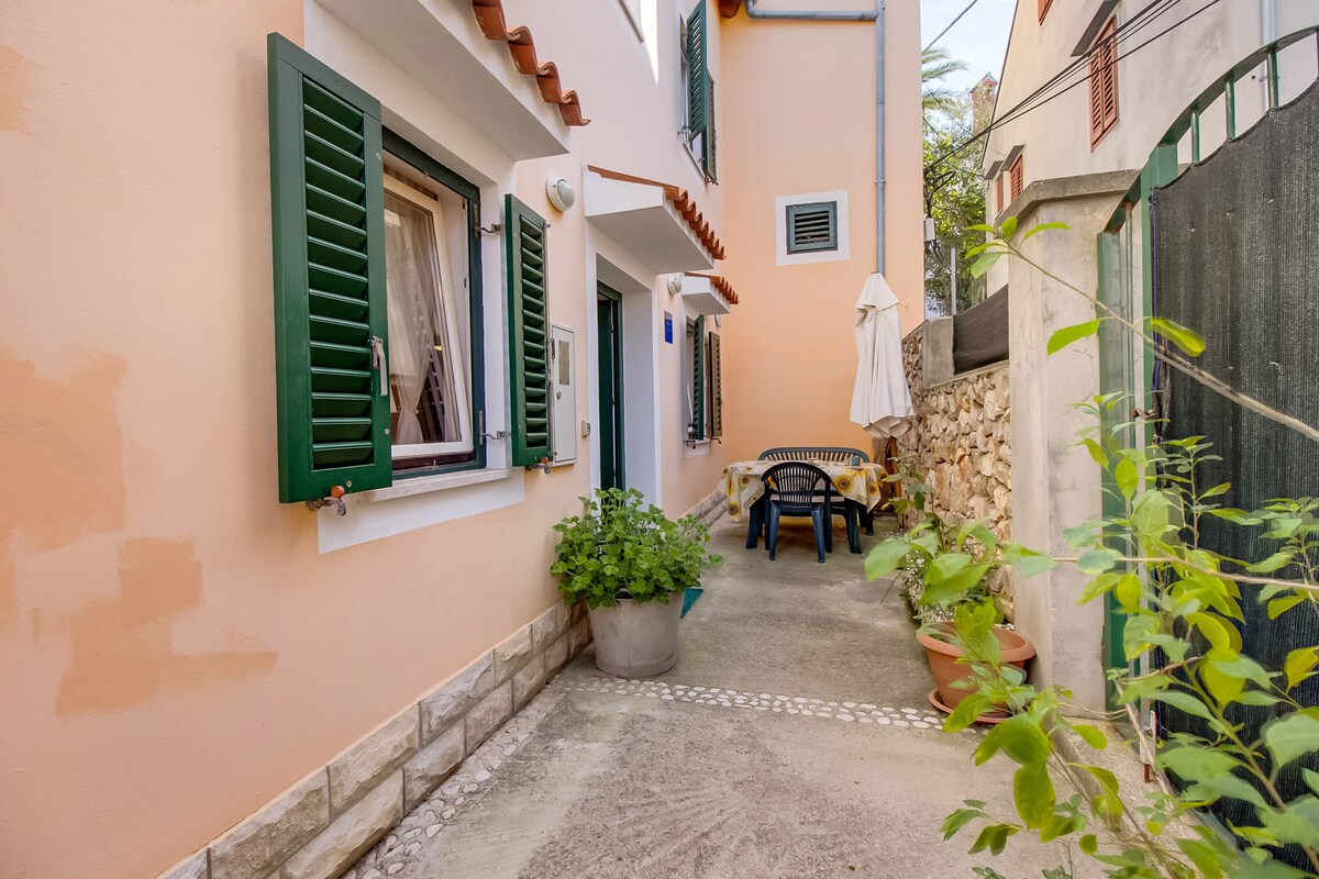 A charming outdoor space is shown, featuring terracotta walls and green shutters. A wooden table and chairs occupy the area, surrounded by potted plants. A white umbrella is propped against the wall, providing shade. The pathway is paved and leads to the entrance of the apartment.