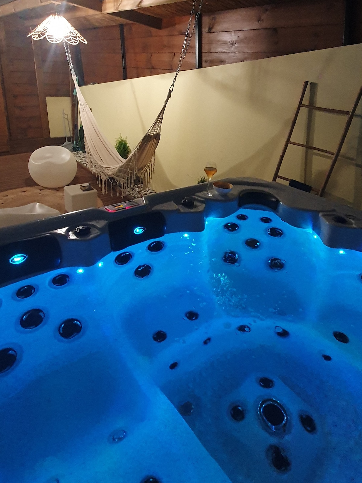 A stylish jacuzzi is illuminated with soft blue lighting, inviting relaxation. A hanging hammock is visible in the background, surrounded by greenery. A decorative ladder leans against the wall, contributing to the overall cozy ambiance.