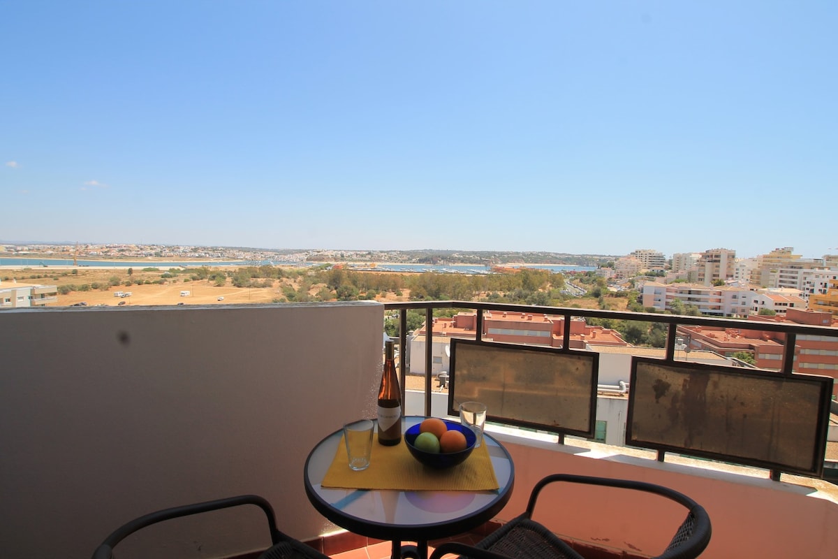 An outdoor balcony offers a view of the surrounding landscape, featuring a combination of hills and urban development. A round table is set with a bottle and two glasses, along with a bowl of colorful decorative balls, inviting relaxation in the open air.