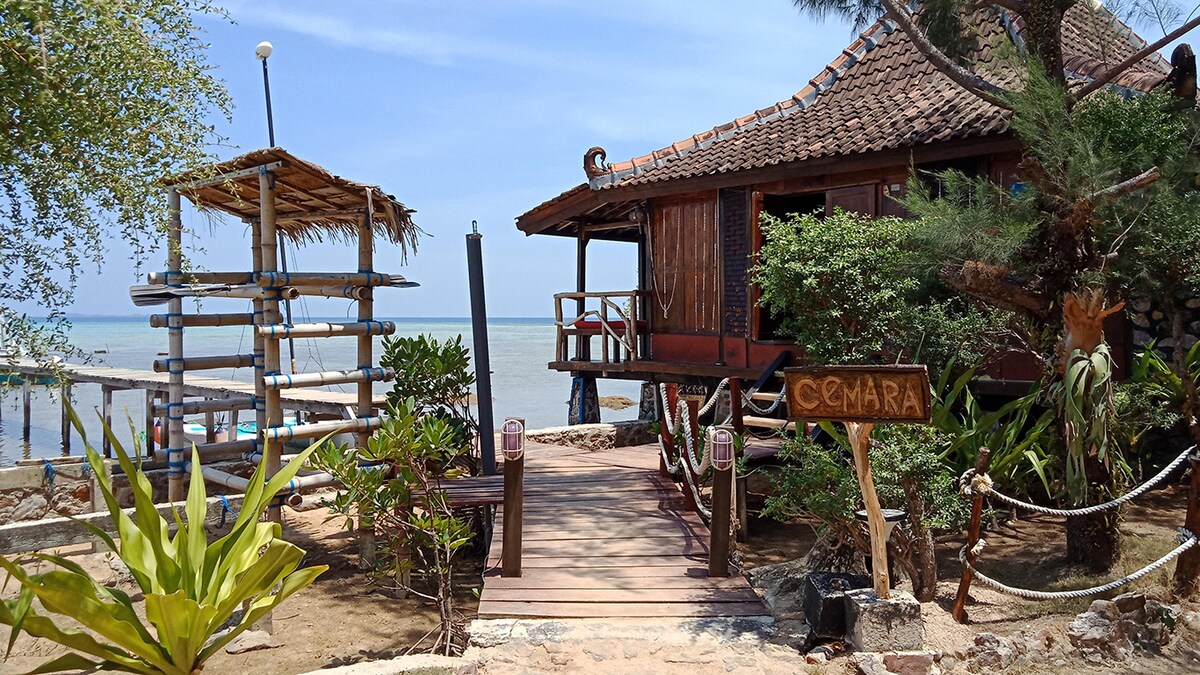 A wooden pathway guides to a traditional house, surrounded by lush greenery and palm trees. A bamboo structure for seating is visible on the left, while the tranquil sea can be seen in the background under a clear blue sky.