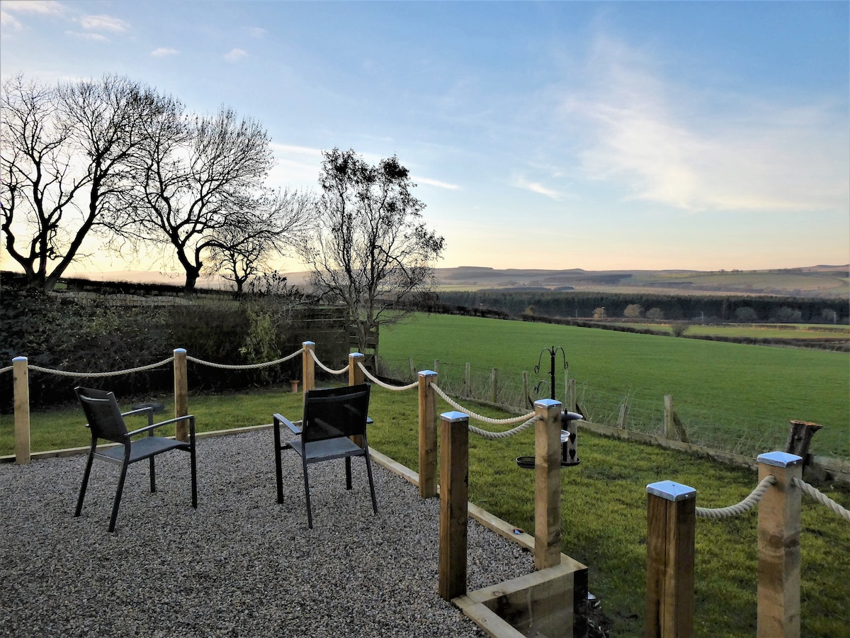 A secluded raised terrace is shown, featuring two black chairs positioned on a gravel surface. The space offers unobstructed views of the lush green fields and rolling hills in the distance, framed by bare trees against a clear sky.