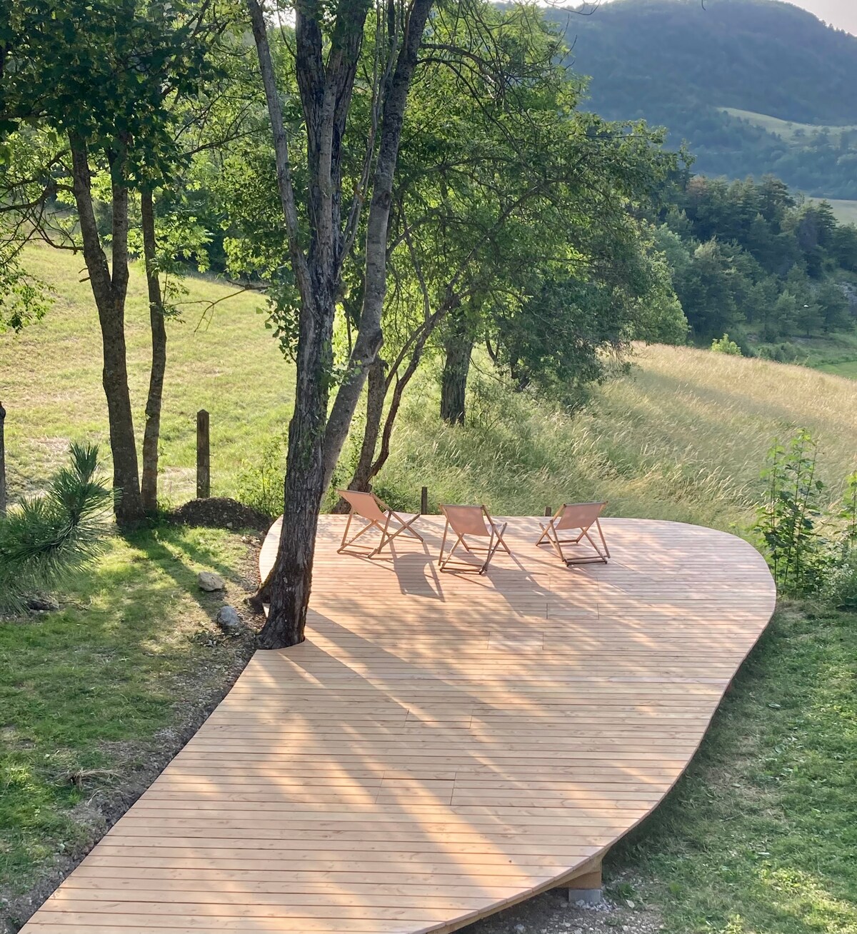 A wooden terrace is set among trees, featuring four light-colored chairs arranged around a small table. The surrounding landscape includes gentle hills and tall grass, offering a serene and natural setting.