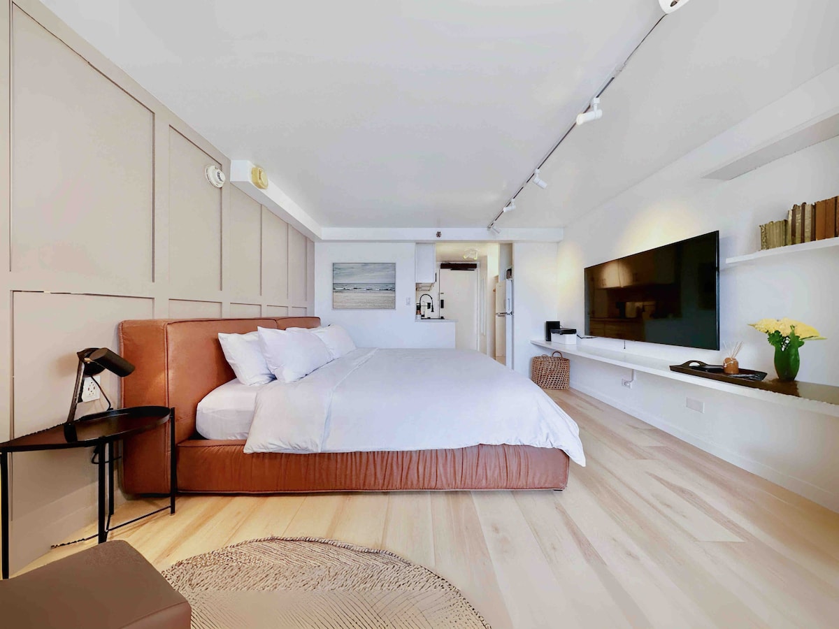 A spacious studio room features a large king bed with fresh linens and a memory foam mattress. A modern flat-screen television is mounted on the wall, accompanied by decorative shelves. Soft lighting is provided by a bedside lamp and natural light enters through a nearby window.