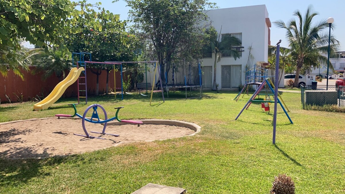 A grassy area features a children's playground with a yellow slide, swings, and a seesaw. Trees provide shade, and a sandy pit is present for play. The background shows a white structure, adding to the open, recreational space.