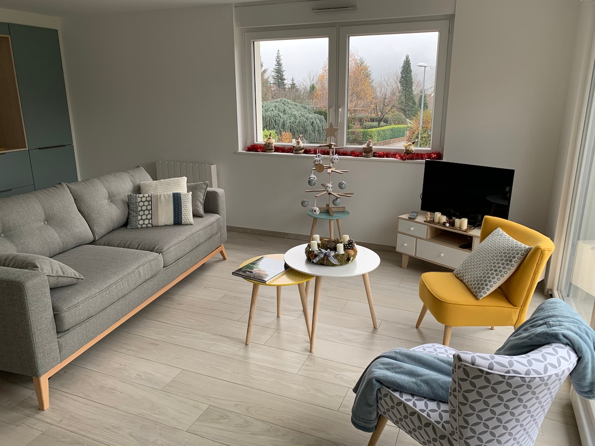 A modern living area is presented with a light grey sofa and a yellow armchair. A white coffee table sits in the center, adorned with decorative items. A large window offers a glimpse of the outdoors, while a small television is positioned nearby.